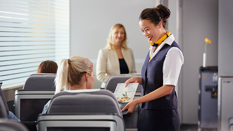 Eine Flugbegleiterin bietet in einer Trainingssituation einer Passagierin die Speisekarte an.