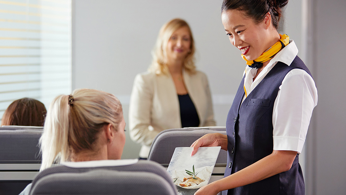 Eine Flugbegleiterin bietet in einer Trainingssituation einer Passagierin die Speisekarte an.