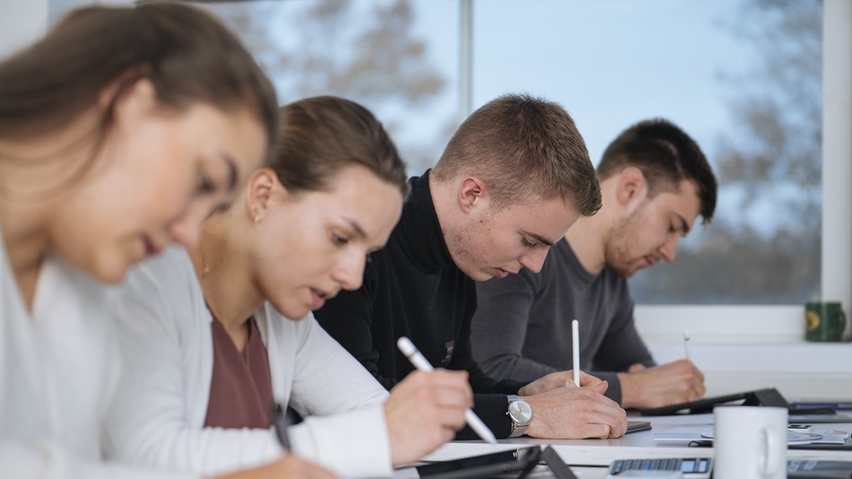 Three people sit one behind the other at tables in front of laptops and solve tasks