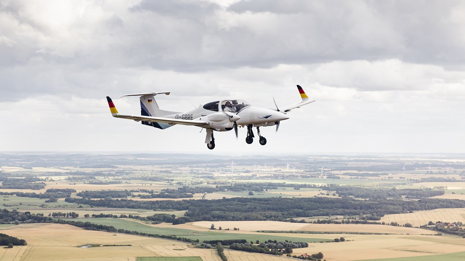 A DA42, with two people visible inside, is flying over fields and meadows under a cloudy sky.